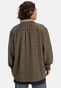 Camisa de cuadros de manga larga en marrón y negro, con un cuello beige, corte relajado y un yugo trasero abotonado para un detalle adicional.