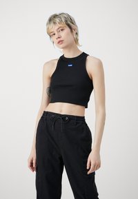 Black cropped tank top in ribbed fabric with a round neckline and thin straps. Features a blue logo tag on the front. Paired with black cargo pants.