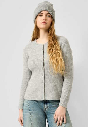 Junge Frau mit langen welligen blonden Haaren, die eine graue Strickmütze, eine graue geknöpfte Strickjacke und blaue Jeans vor einem schlichten Hintergrund trägt.