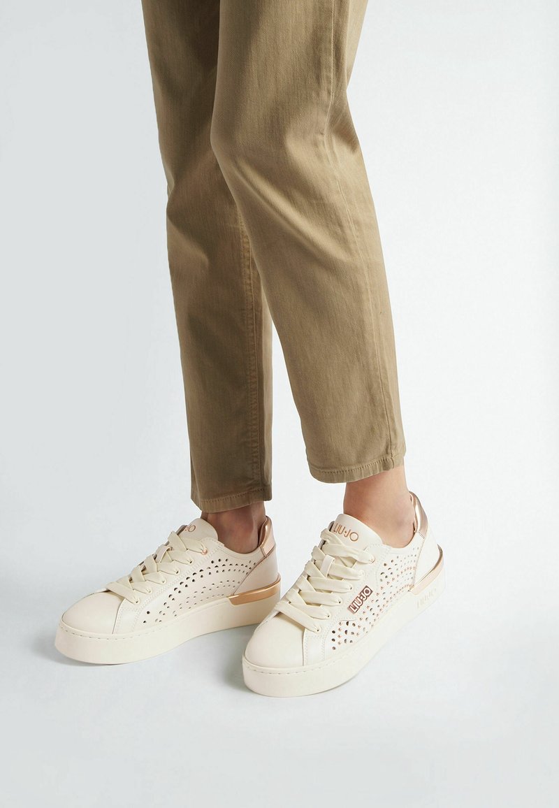 Personne portant un pantalon court beige et des baskets blanches avec des côtés perforés et des accents en or rose, debout sur un fond clair.