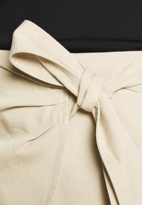 Gros plan sur un tissu beige noué à la taille au-dessus d'un vêtement noir à côtes, mettant en valeur la texture et les détails de couture.