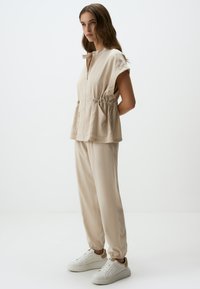 Completo beige in due pezzi, composto da un top a maniche corte con collo a zip e vita regolabile con cordino, abbinato a pantaloni larghi e sneaker bianche.