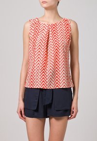 Top senza maniche di color crema con un motivo a zigzag rosso, caratterizzato da un frontale plissettato e una vestibilità ampia. Abbinato a pantaloni corti blu navy. Tessuto testurizzato.