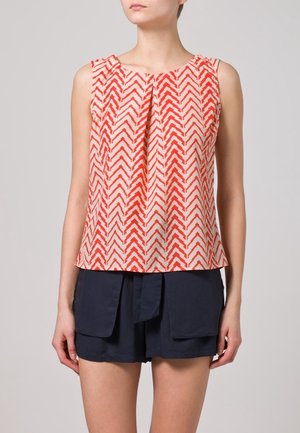 Débardeur sans manches crème avec un motif en zigzag rouge, présentant un devant plissé et une coupe ample. Associé à un short marine. Tissu texturé.