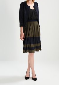 Veste bleu marine, blouse noire, jupe plissée vert olive et bleu marine avec des rayures horizontales, associées à des talons hauts noirs.