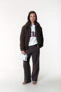Brun fluffy jakke med høy krage, hvit genser med "REBEL" tekst, løse mørke bukser og en liten hvit håndveske med gulldetaljer.