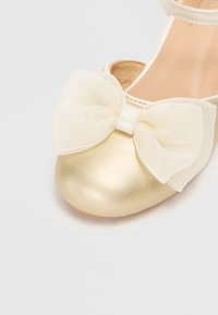Gullgule syntetiske ballerinasko med rund tå og en stor kremfarget sløyfe foran, med en delikat tekstur og ankelstropp.