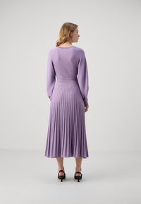 Robe lilas à côtes avec manches longues, taille cintrée et jupe plissée. Texture lisse et design longueur cheville. Sandales à talons noires.