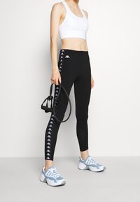 Czarne legginsy z białym logo Kappa i wzorzystą boczną taśmą, w połączeniu z dopasowanym białym topem i niebieskimi butami sportowymi.