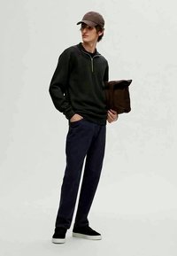 Pullover verde escuro com gola de zip, calças azul-escuras, sapatilhas pretas e uma mala castanha. O modelo usa um boné castanho e está em pé de forma descontraída.