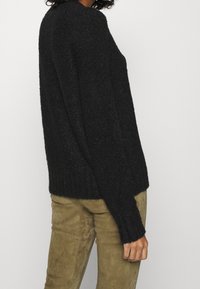 Pull en maille noire texturée avec manches longues et ourlet côtelé, porté avec un pantalon ajusté de couleur marron clair, aspect daim.