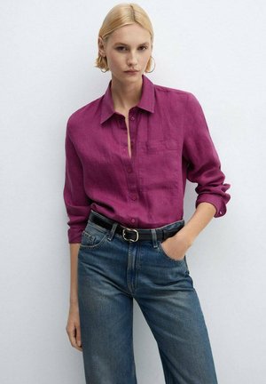 Blonde woman wearing a magenta button-up shirt, blue jeans with a black belt, and gold hoop earrings, standing against a white wall.
