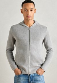 Lys grå hettejakke med et strukturert strikkemønster, som har hette og lange ermer, kombinert med blå denimjeans.