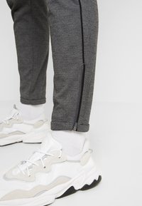 Jambes inférieures portant un pantalon gris à fermetures éclair latérales et des baskets blanches avec des chaussettes blanches sur un fond clair et uni.