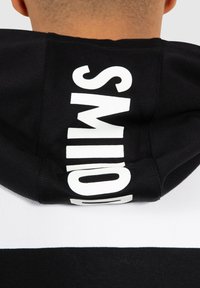 Sudadera en blanco y negro que presenta un gran y audaz texto "SMILO" en la parte trasera. El material parece suave y tiene una textura lisa.