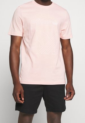 Mann trägt hellrosa Adidas-T-Shirt mit weißem Logo und schwarzen Shorts, steht vor schlichtem hellem Hintergrund.