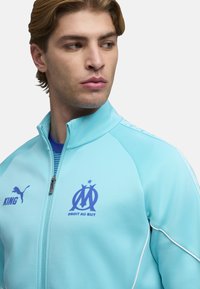 Chaqueta azul claro con cremallera, con gráficos azules que incluyen "KING", un logotipo y "DROIT AU BUT". Presenta una textura suave y un diseño ajustado.