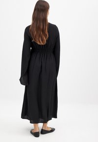 Sort langærmet kjole lavet af let, struktureret stof med en samlet talje, der når til midt på læggen, parret med enkle sorte flade sko.