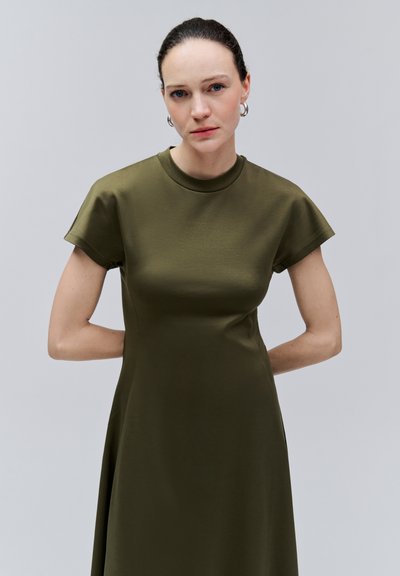 Femme aux cheveux tirés en arrière portant des boucles d'oreilles créoles et une robe à manches courtes vert olive, debout les mains derrière le dos.