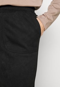 Mustat elastiset vyötäröhousut, jotka on valmistettu pehmeästä ja teksturoidusta materiaalista, edessä tasku ja näkyvä ompelu. Yhdistetty vaaleanruskean yläosan kanssa.