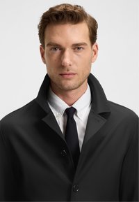 Manteau noir à col haut, avec fermeture à boutons. Porté sur une chemise blanche et une cravate sombre, le tissu semble lisse et bien coupé.