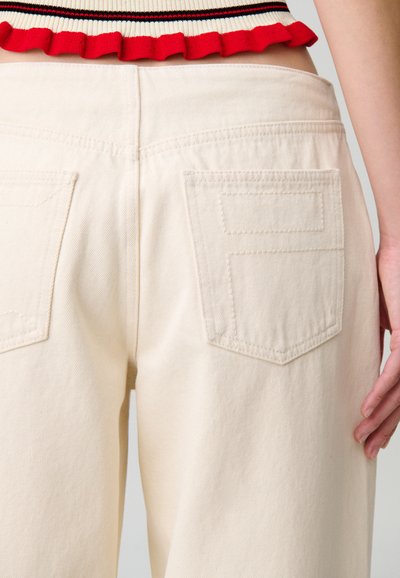 Pantalones de algodón beige con dos bolsillos traseros, cintura alta y textura suave. La cintura es visible junto a un top corto de contraste.