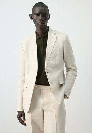 Uomo che indossa un completo beige chiaro con pantaloni abbinati e una camicia verde oliva scuro con colletto, in piedi davanti a uno sfondo chiaro e uniforme.