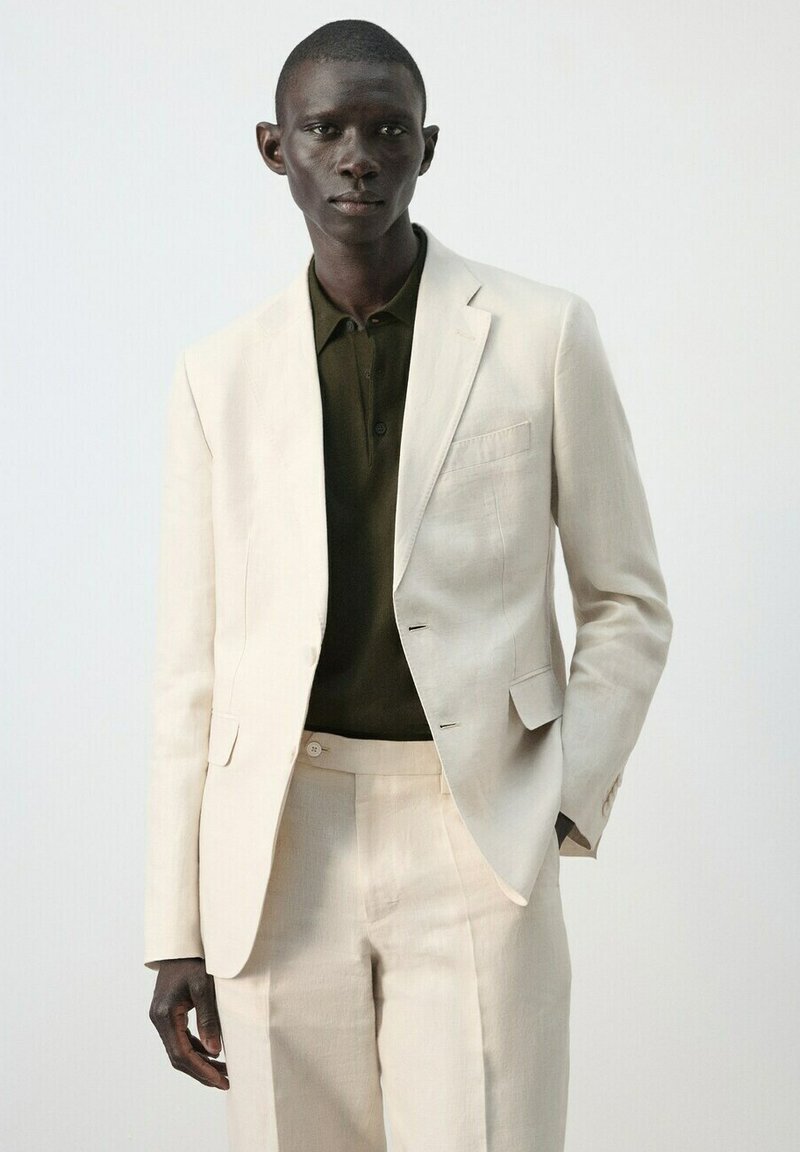 Muškarac u svijetlosmeđem odijelu s odgovarajućim hlačama i tamno maslinastozelenoj košulji s ovratnikom, stoji na jednostavnoj svijetloj pozadini.