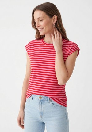 Mujer con cabello castaño hasta los hombros, vestida con una camiseta sin mangas a rayas rojas y rosas y vaqueros azul claro, sonriendo y tocándose el cuello.