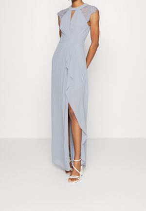 Kvinde iført lyseblå, ærmeløs kjole med blonder på kasketærmerne, nøglehul foran, flæset slid på skørtet og hvide sandaler med stropper.