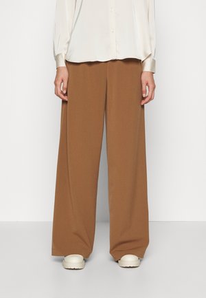 Person iført løstsiddende brune bukser med brede ben, en cremefarvet bluse med lange ærmer og knappede manchetter samt hvide sko med tyk sål.