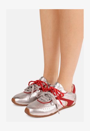 Piernas con zapatillas plateadas y rojas de cordones con suelas de goma, con cordones mezclados en rojo y gris atados de forma suelta.