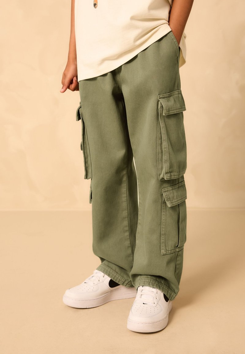 Enfant portant un pantalon cargo vert olive avec de grandes poches latérales et des baskets blanches, debout avec une main dans la poche.