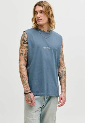 Młody mężczyzna z długimi włosami, ubrany w niebieską koszulkę bez rękawów z napisem „JJ Studios”, jasne dżinsy oraz z licznymi tatuażami na ramionach i dłoniach.