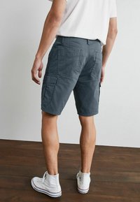Graue Cargo-Shorts mit mehreren Taschen, lockerer Passform und glatter Textur, kombiniert mit weißen High-Top-Sneakern auf einem Holzboden.