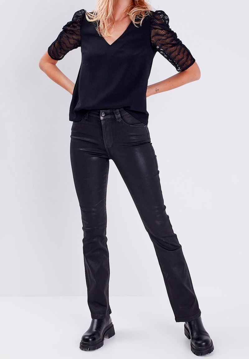 Femme portant un haut noir à col en V avec des manches bouffantes en dentelle, un pantalon noir ajusté et des bottes noires épaisses, debout les mains sur les hanches.