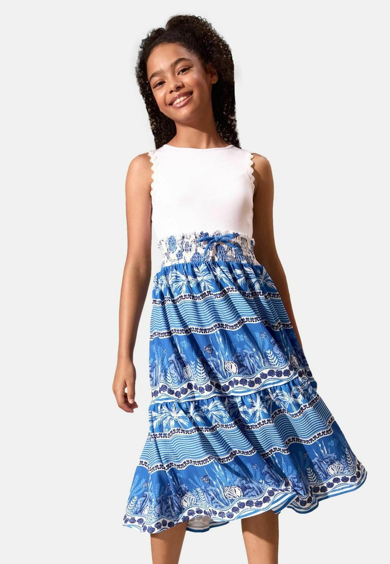 Fille souriante aux cheveux bouclés portant un haut blanc sans manches et une jupe bleu à volants à motifs sur un fond uni.