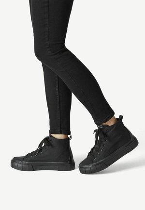 Musta kangaskorkeavartinen lenkkari, jossa on teksturoidut kumipohjat, nauhakiinnitys ja takana vetotappi. Yhdistetty mustiin kapeisiin farkkuihin.