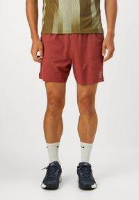 Bordeaux idrettsshorts med flat midje, sidelommer og liten logo-detalj. Brukt med en mønstret skjorte og sportssko.