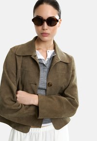 Oliwkowa zamszowa kurtka z kołnierzem, zapięciem na guziki i rękawami do trzech czwartych, zestawiona z szarym polo i okularami z brązowymi oprawkami.