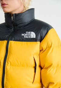 Dzeltena pūkainā jaka ar melnu augšējo daļu, kas aprīkota ar augstu apkakli, priekšējo rāvējslēdzēju, sānu kabatu un baltu logo uz krūtīm.