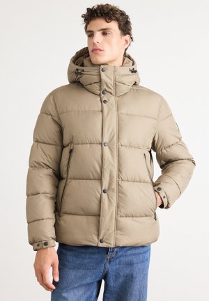 Doudoune beige avec capuche, présentant un design cousu, une fermeture à boutons-pressions et des poches latérales zippées, associée à un jean bleu.