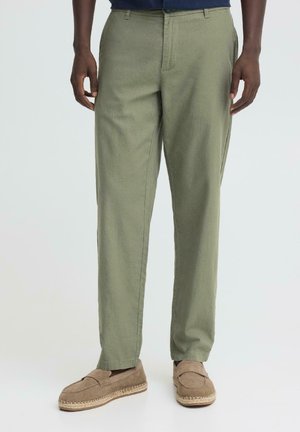 Man wearing olive green straight-leg pants and beige suede slip-on shoes, standing with hands relaxed at sides.