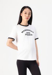 Hvit bomull t-skjorte med svart ringer rundt halsen og ermer. Har "MARSEILLE 1975 FRANKRIKE" trykt i en fet buet design.