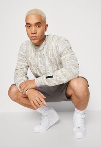 Light gray textured sweater with intricate patterns, paired with gray shorts and white high-top sneakers. The individual poses in a squat.