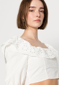Top cropped branco com um grande colarinho de renda bordada, mangas curtas, textura de algodão suave e design ajustado, exibindo padrões florais abertos.