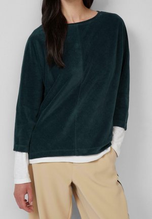 Femme portant un haut en velours côtelé vert foncé à manches longues superposé à une chemise blanche, associé à un pantalon beige, debout devant un fond gris.