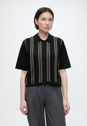 Ung person med kort sort hår iført en sort kortærmet skjorte med lodrette olivengrønne striber og grå bukser.