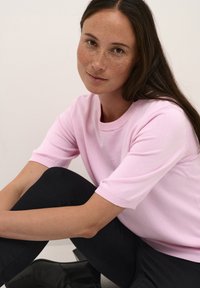 Svetlý ružový sveter s krátkymi rukávmi a rebrovaným okrúhlym výstrihom, nosený s čiernymi nohavicami. Hladká textúra, elastický strih. Minimalistický štýl.