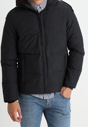 Mężczyzna w ciemnoszarej pikowanej kurtce na zamek, pod którą ma koszulę w kratę, oraz niebieskich dżinsach, z jedną ręką w kieszeni.